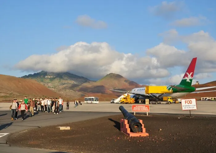 Passengers walking on an airstrip beside a plane with mountains in the background at a remote location.