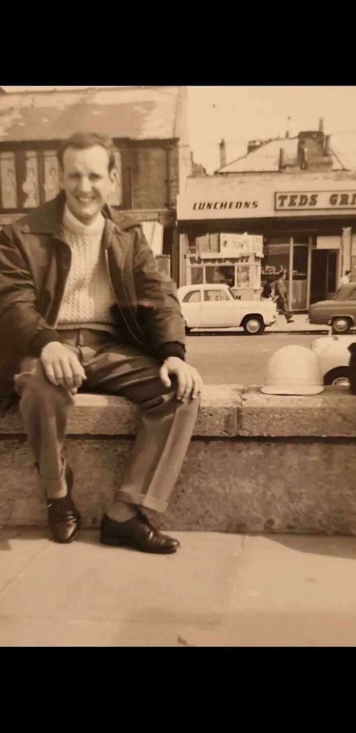 Vintage black and white photo of a man sitting outdoors in front of a diner, classic cars in background, where was this taken post.