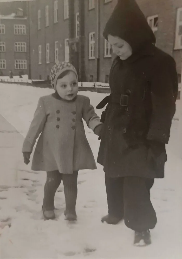 Two children holding hands walking in snow, wearing winter coats and hats, capturing a vintage where was this taken moment.