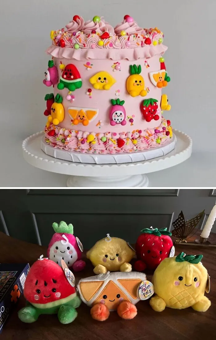 Pink frosted cake decorated with colorful fruit-themed designs and matching plush fruit toys on a wooden table.