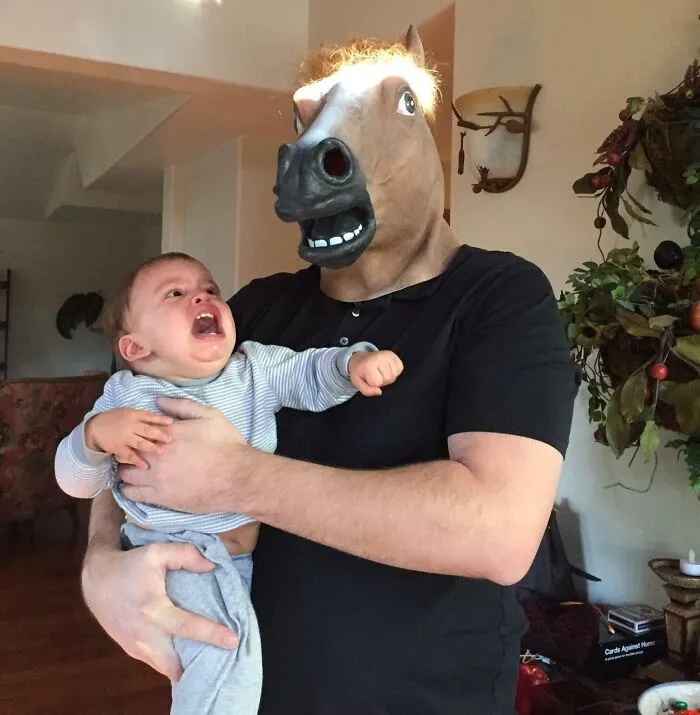 Man wearing a horse mask holding a crying baby, showcasing playful and wholesome moments men never grow up from new pics.