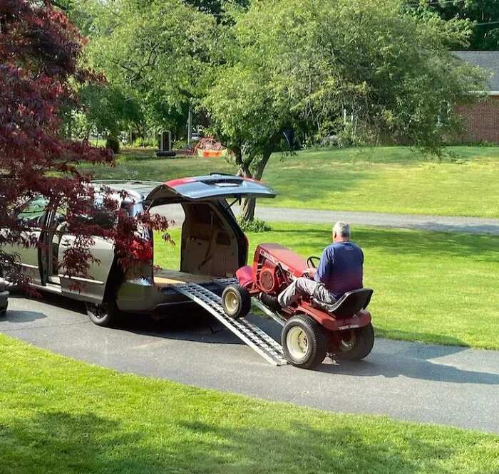 Man driving riding lawn mower onto van ramp in a green yard, showing men never grow up in wholesome moment.