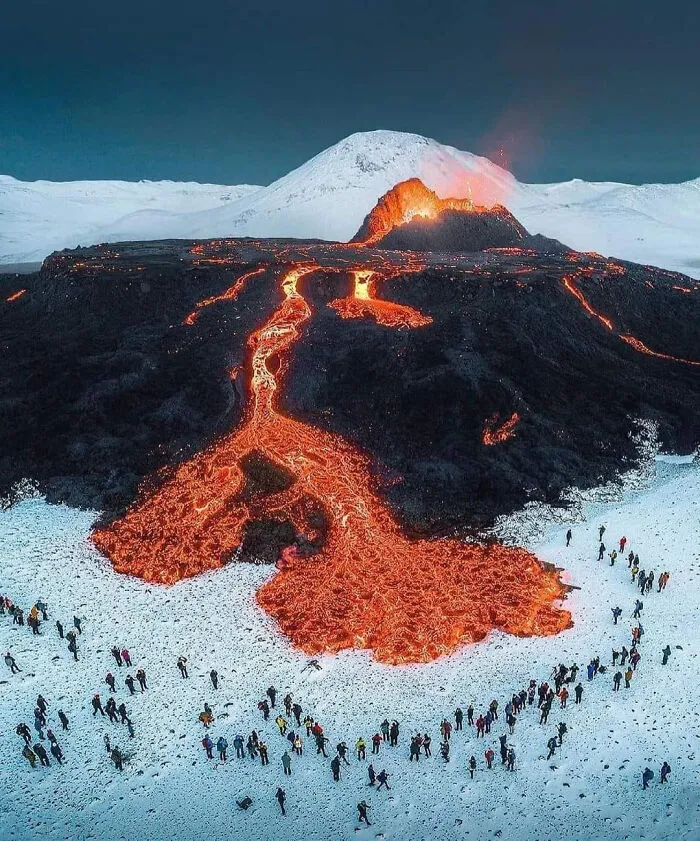 A volcanic eruption with flowing lava surrounded by snowy landscape and many people observing, exploring existence and reality.