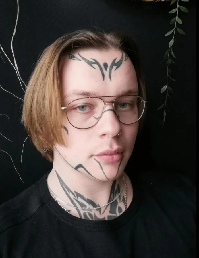 Young man with fierceflow and beautiful hair wearing glasses and tribal face and neck tattoos against a dark background