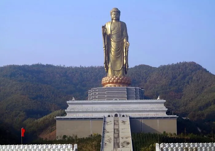 Golden standing Buddha sculpture on pedestal with mountainous background, a spectacular sculpture attracting global travelers.