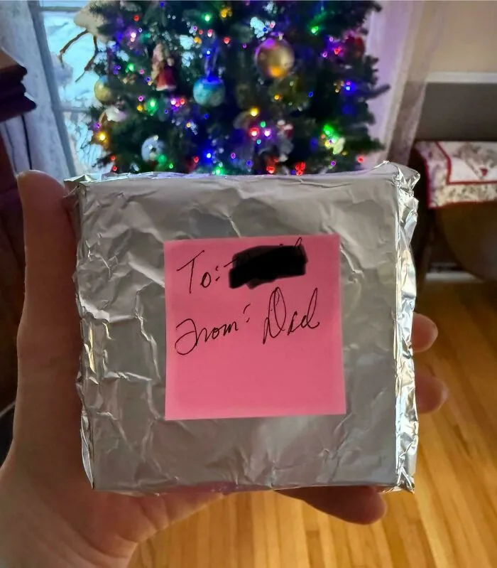 Hand holding a foil-wrapped gift with a pink note from Dad in front of a decorated Christmas tree, showing wholesome moments.