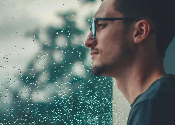 Young man wearing glasses looking thoughtfully out a rain-covered window, inspired by genius cleaning hacks for laziness.