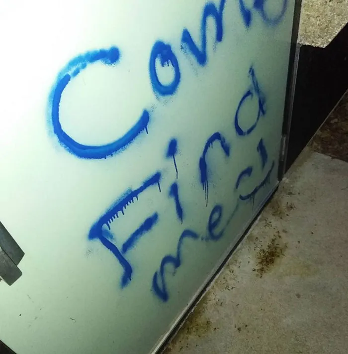 Blue graffiti spelling creepy message on a white door inside a dark urban exploration site at night.