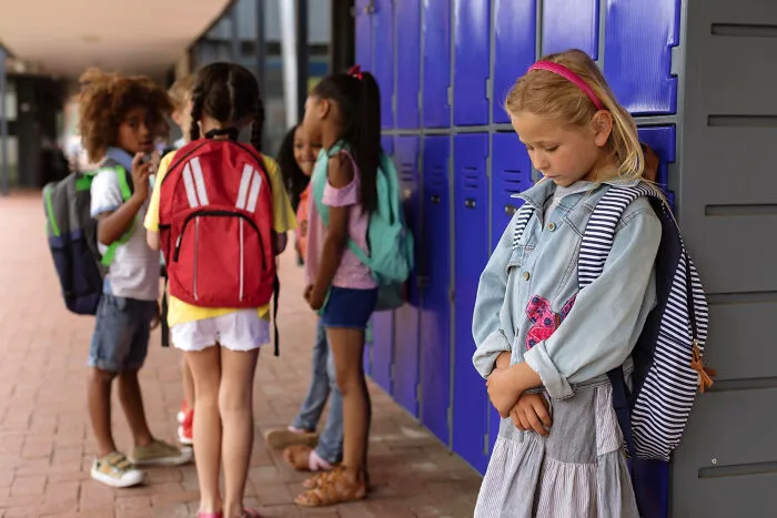 A young girl looking sad and isolated by school lockers while other kids with backpacks socialize nearby, illustrating a backfired lie.