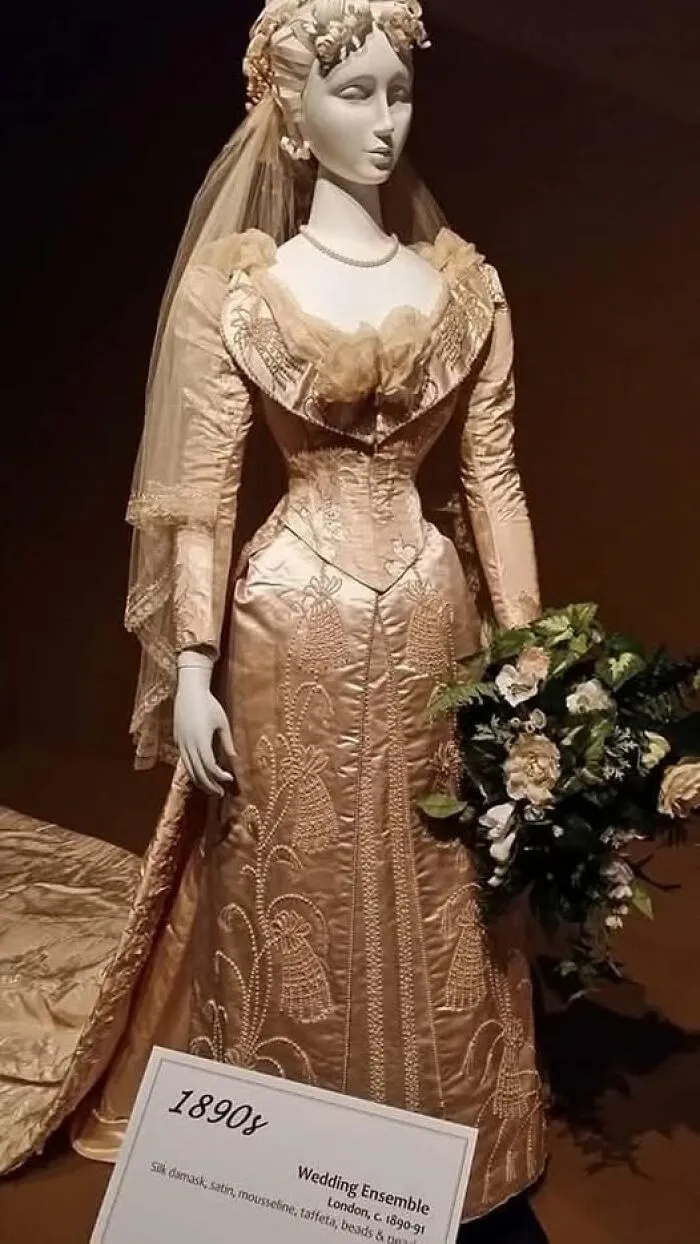 Mannequin dressed in a Victorian-Edwardian wedding ensemble from the 1890s holding a bouquet of flowers.