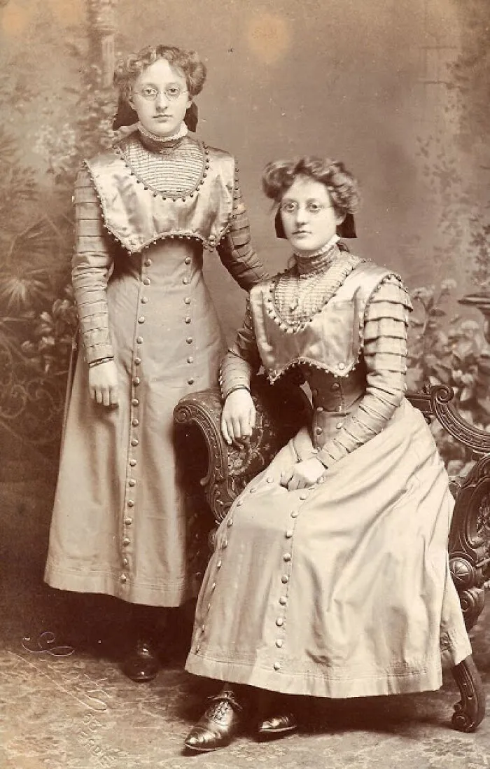 Two women in Victorian-Edwardian dresses with high collars and buttons posing in a vintage indoor setting.