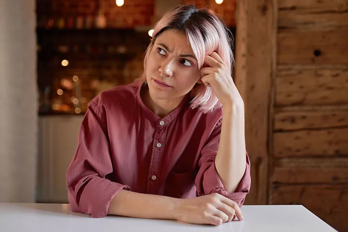 Young woman with pink hair and nose ring looks confused and thoughtful while sitting at a table indoors.