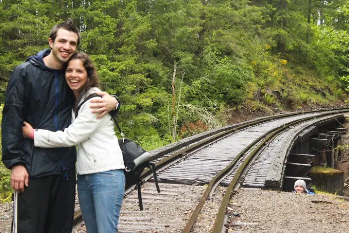 Pareja sonriente abrazándose cerca de vías de tren con cameo inesperado asomando detrás, fotos arruinadas divertidas.