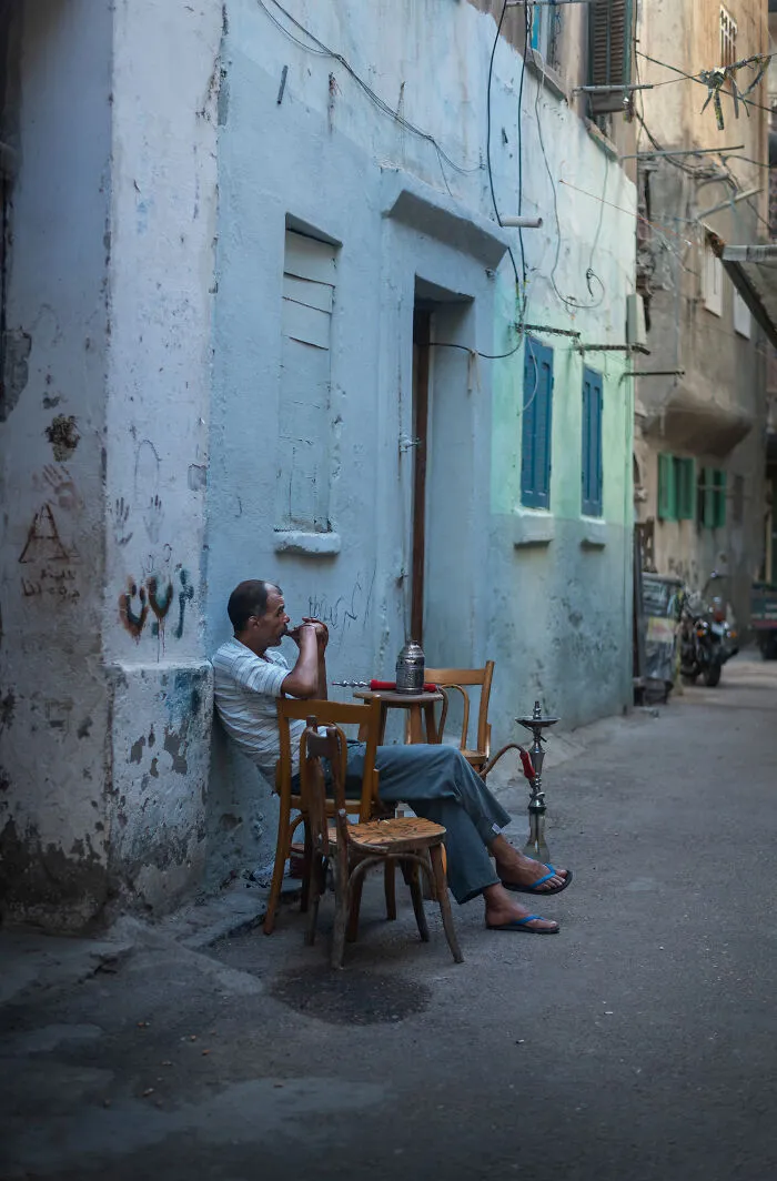 Man smoking hookah while sitting on wooden chairs in a narrow alley, captured in street photography around the world.