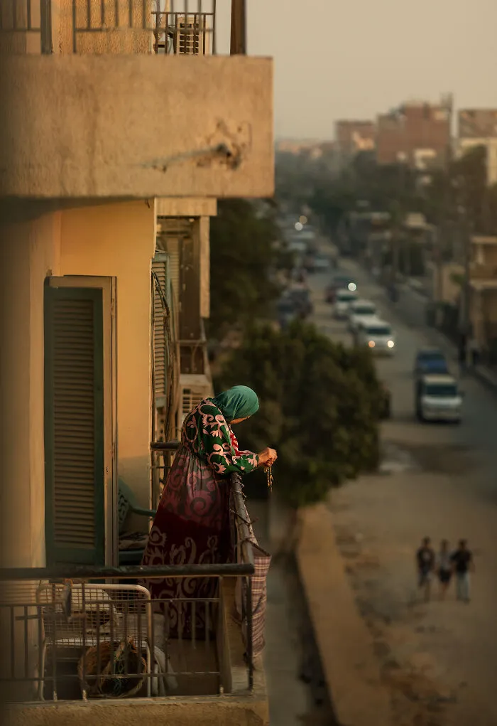 Woman in colorful dress and headscarf leaning on balcony railing overlooking busy street in street photography shot