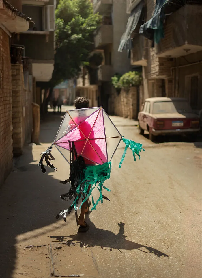 Child walking in a sunlit street holding a colorful kite, captured in a candid street photography shot.
