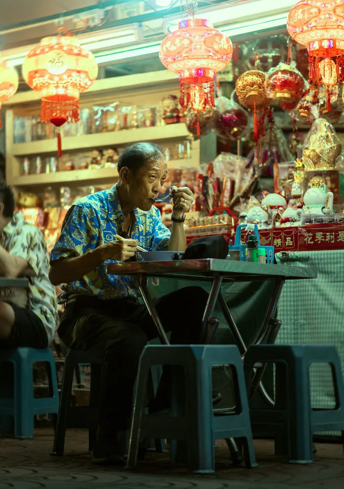 Street photography capturing a man eating at a vibrant night market with colorful lanterns and traditional decorations.