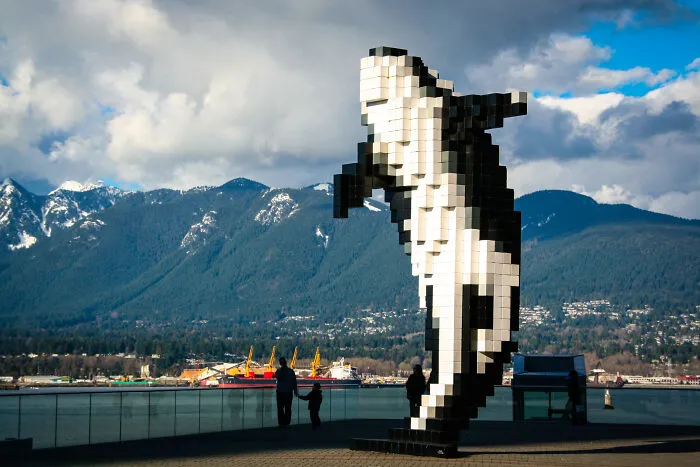 Pixelated killer whale sculpture near waterfront with people and mountains in the background, a spectacular sculpture for travel.