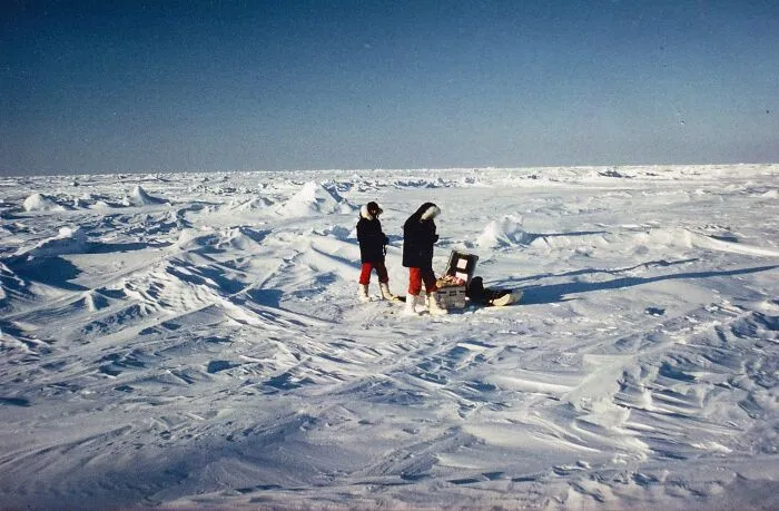 Two people in cold weather gear exploring a vast snowy remote location with icy terrain and clear blue sky.