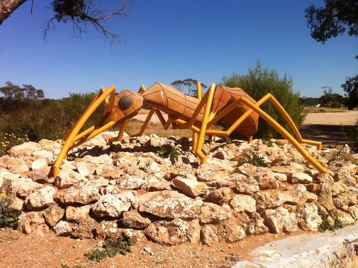 Large spectacular ant sculpture made of wood and metal displayed outdoors on rocky terrain under clear blue sky.