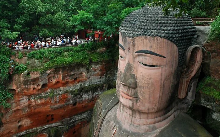Massive ancient Buddha sculpture carved into red cliff surrounded by greenery and tourists, a spectacular global sculpture.