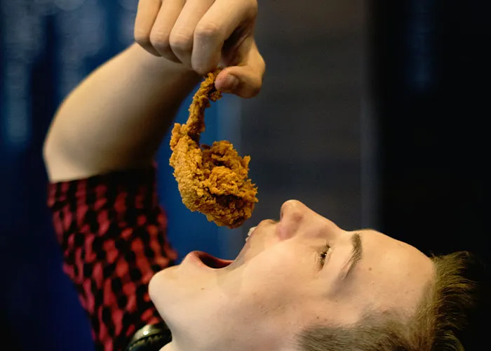 Person in a red and black shirt holding fried chicken above their open mouth in a chaotic food moment witnessed by many.