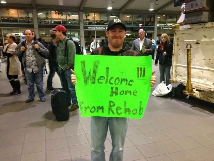 Man holding bright green welcome home sign at airport, showing wholesome moments that prove men never grow up.
