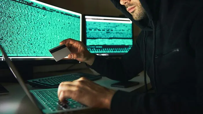 Person in a dark hoodie holding a credit card and typing on a laptop with multiple screens showing code, illustrating scams.