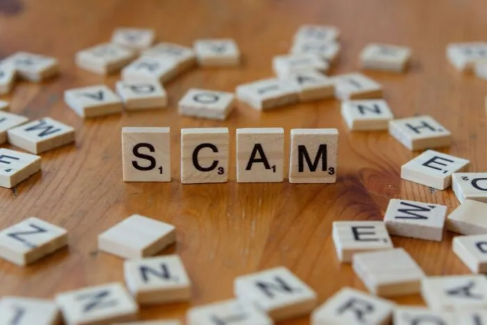 Scrabble tiles arranged on a wooden table spelling scam, surrounded by scattered letter tiles representing people falling for scams.