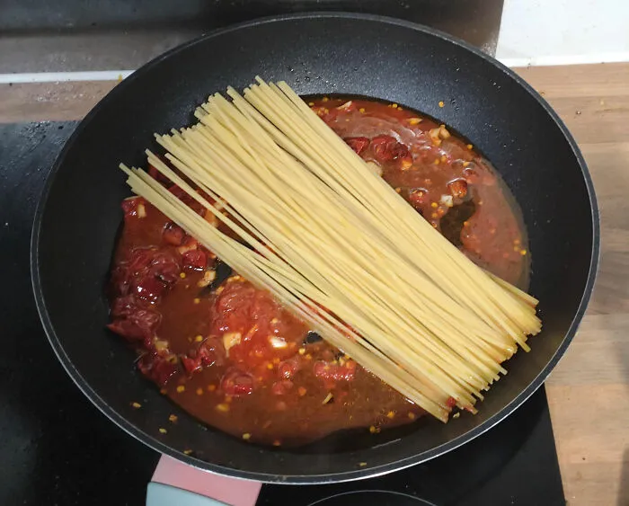 Uncooked spaghetti placed over a pan with tomato sauce and seasonings, demonstrating kitchen tips and cooking skills.