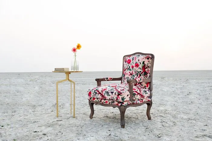 Vintage floral armchair and small table with flowers and books placed alone on a barren ground, evoking bizarre and creepy park scenes.