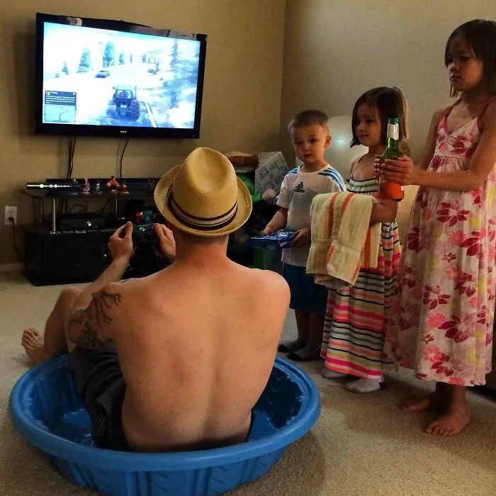 Man playing video games sitting in a plastic pool indoors, surrounded by three children holding toys and towels, wholesome moments.