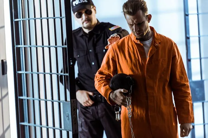 Prisoner in an orange jumpsuit holding a ball and chain as a police officer opens a jail cell door behind him.