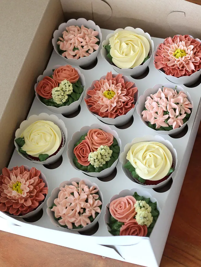Box of baked goods decorated with intricate frosting flowers in various shades of pink, cream, and green leaves.