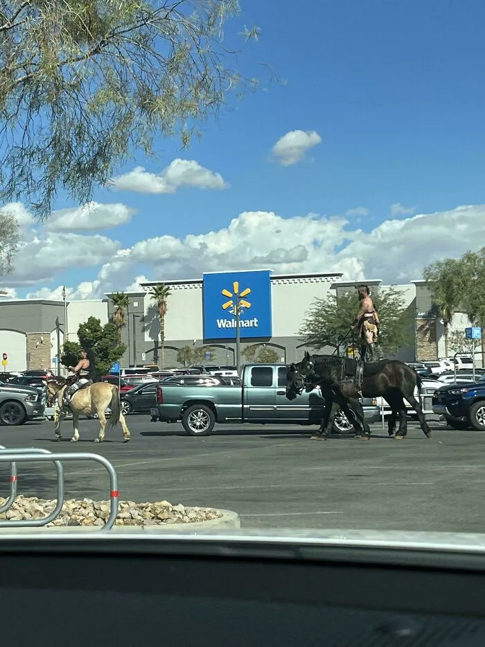 Two people riding horses in a Walmart parking lot on a clear day with blue skies and scattered clouds.