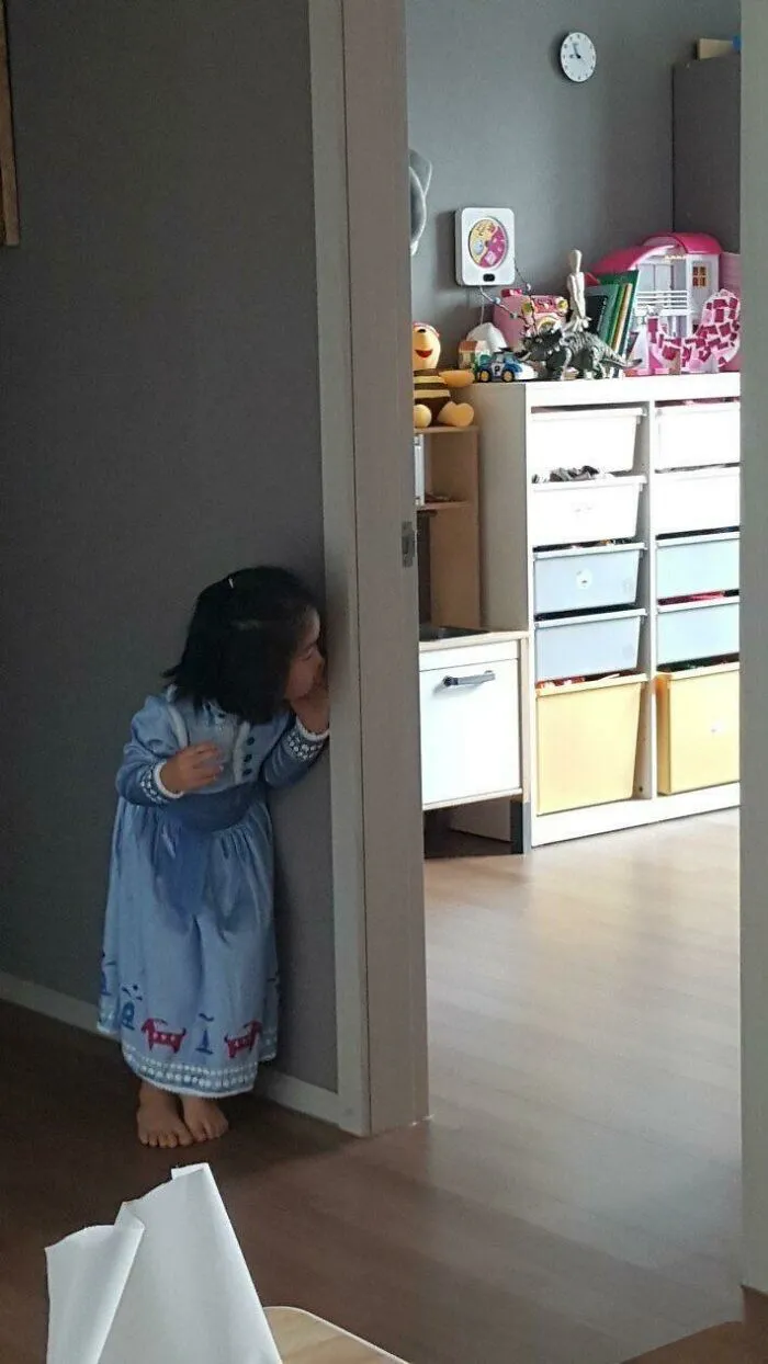 Little girl in blue dress peeking around a door in a cozy room with toys and storage, heartwarming photo moment.