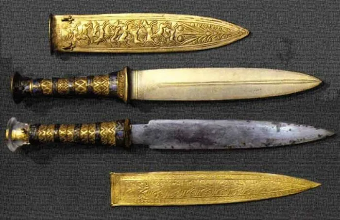 Two ancient Egyptian ceremonial daggers with ornately decorated golden sheaths displayed on a dark background.