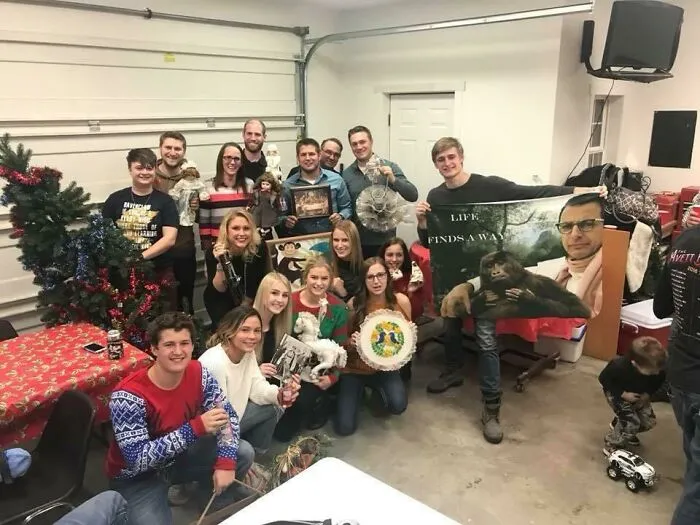 Group of people in a garage holding bizarre Christmas gifts including unusual decorations, photos, and toys.