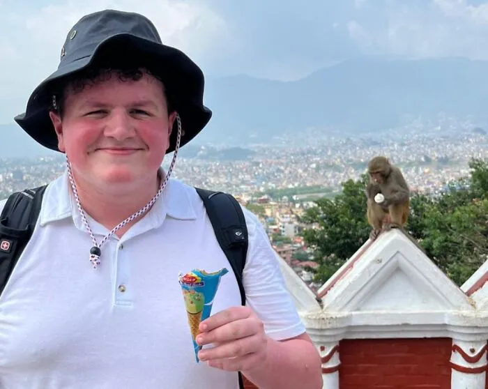A sneaky pet monkey trying to steal food from a man holding an ice cream cone outdoors with a cityscape background.