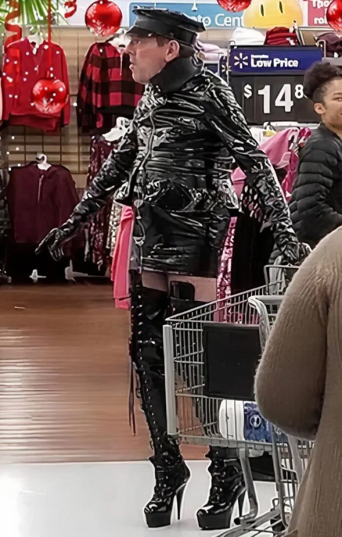 Person in shiny black leather outfit and high heels pushing a cart inside Walmart, dressed unusually for the store environment.