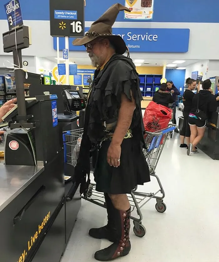 Man in wizard hat and black costume standing at Walmart checkout with shopping cart behind him in store aisle.