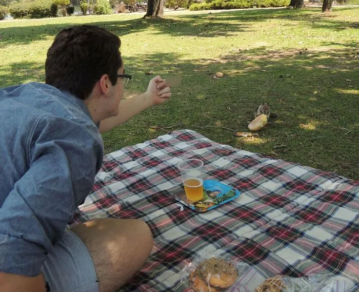 Man on picnic blanket in park caught a sneaky pet squirrel trying to steal food during an outdoor snack moment.