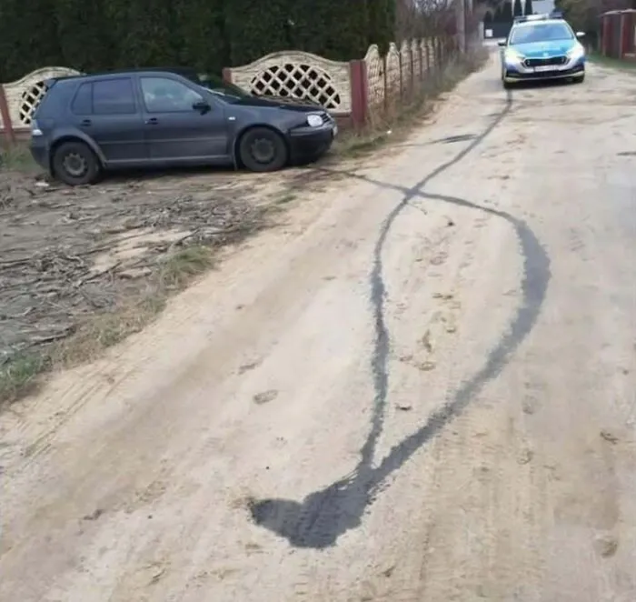 Tire skid marks on a dirt road creating a humorous shape in a rural Eastern Europe setting with parked cars nearby.