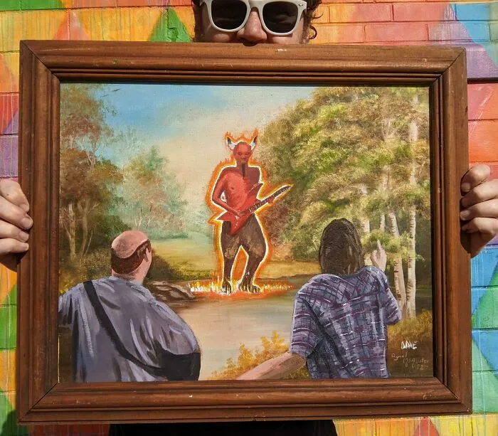 Person holding a personalized thrifted painting featuring a devil playing guitar in a forest, adding something funny or unexpected.
