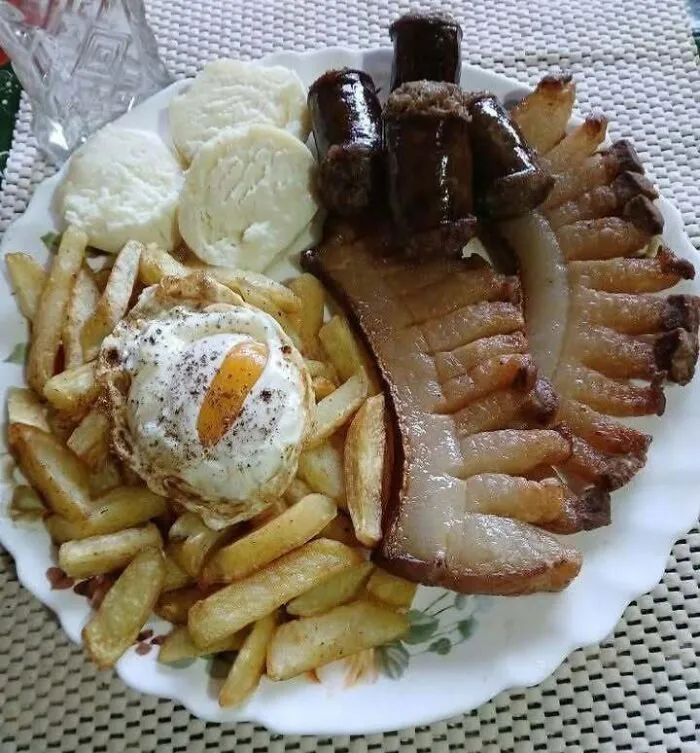 Traditional Eastern European meal with fried egg, sausages, bacon, potato fries, and dumplings on a white plate.