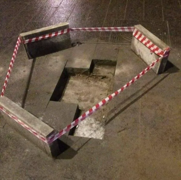 Damaged sidewalk hole surrounded by concrete blocks and red-and-white caution tape in an urban Eastern Europe setting.