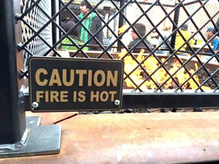 Caution sign reading fire is hot on a metal fence with people and a fire burning behind, funny memes concept.