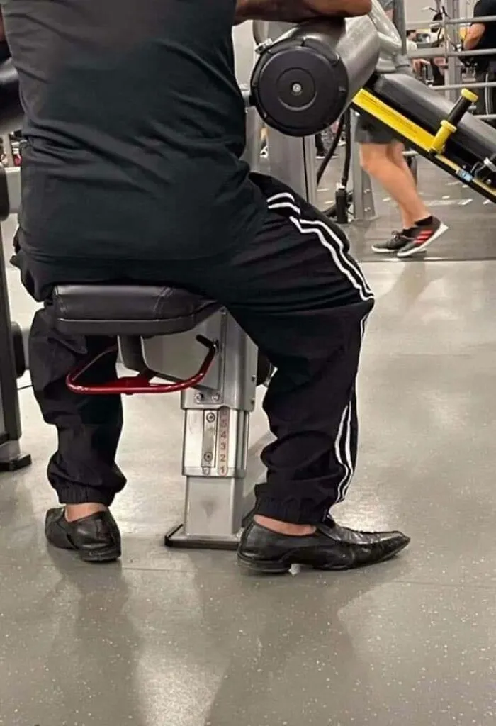 Man in gym wearing black Adidas pants and dress shoes, humorously blending formal and athletic styles, Eastern Europe meme style.