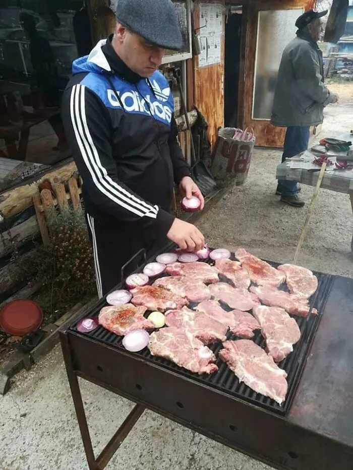 Man in Adidas jacket grilling meat and onions outdoors, capturing an Eastern Europe nostalgic barbecue moment.
