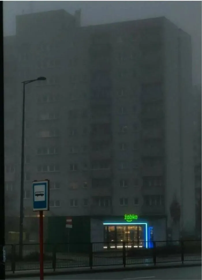 Dimly lit Eastern Europe apartment block with a neon-lit Żabka convenience store in foggy urban setting at dusk